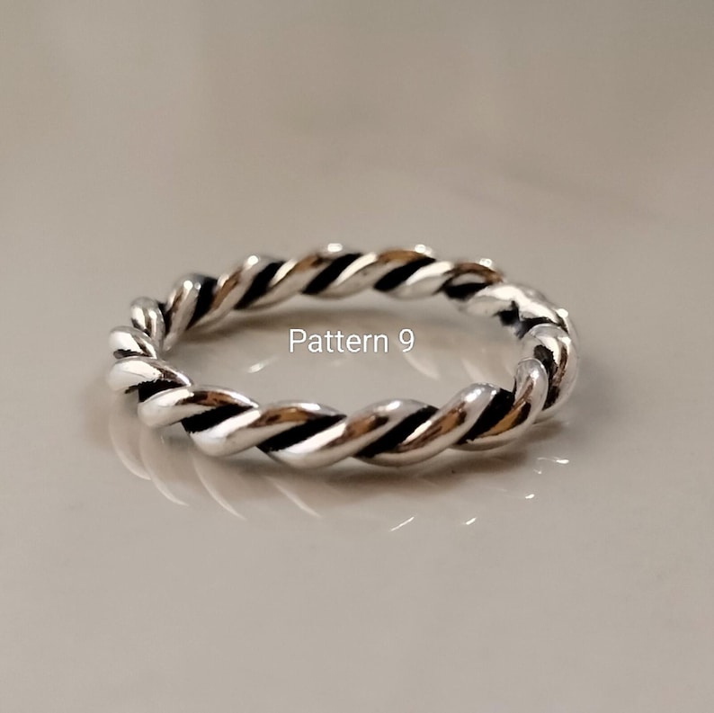 May include: A silver-toned ring featuring a twisted rope design with dark oxidised grooves between the metallic strands. The text Pattern 9 is printed in white in the centre of the ring, which has a polished finish and a continuous circular band.