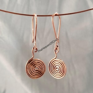 Könnte beinhalten: Ein Paar kupferfarbene Drahtohrringe mit Spiraldesign, die an langen, gebogenen Ohrhaken hängen. Die Metallkomponenten sind zu kreisförmigen Spiralen geformt und der Text TheHeavenOfCraft ist in der Mitte des Bildes deutlich zu erkennen.