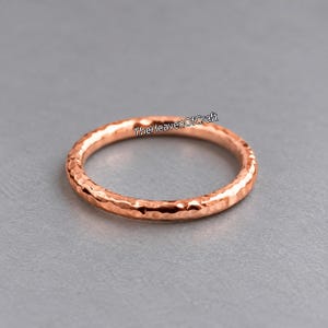 Può includere: Un anello color oro rosa con una superficie martellata e testurizzata. La fascia presenta una finitura continua e irregolare lungo tutta la circonferenza. Il testo TheHeavenOfCraft è visibile in nero sopra l'anello.