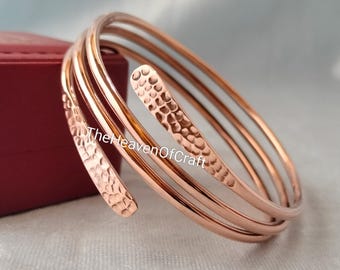 May include: A multi-coil copper bracelet featuring a polished finish and a hammered texture on the tapered ends. The metal wire wraps around in a spiral design. The text TheHeavenOfCraft is visible across the center of the copper bracelet.