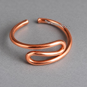 May include: A polished copper ring featuring an open, bypass-style design with a smooth, rounded wire construction. The metal has a warm, metallic orange-brown hue and a reflective finish. The text TheHeavenOfCraft is printed on the ring surface.