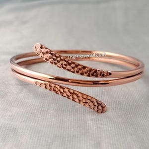 Peut inclure: Un bracelet de type bypass couleur or rose avec un design à double enroulement. Les deux extrémités effilées du bracelet présentent une finition martelée texturée, tandis que la partie centrale reste lisse. Le texte TheHeavenOfCraft est visible.