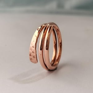 Puede incluir: Un anillo tipo bypass en tono oro rosa con diseño de envoltura múltiple. El anillo consta de dos bandas redondeadas y lisas, y una banda plana más ancha con un acabado martillado y texturizado que termina en punta.