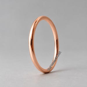 Peut inclure: Une bague en or rose lisse et polie avec un anneau fin et arrondi. La surface du métal reflète la lumière, créant un éclat métallique chaud. Le texte TheHeavenOfCraft est imprimé en petites lettres noires sans empattement sur le côté.