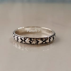 May include: A silver-toned metal ring featuring a repeating embossed pattern of hearts and small floral shapes around the band. The text Pattern 7 is printed in white in the centre of the ring. The metal has a polished finish with dark accents.