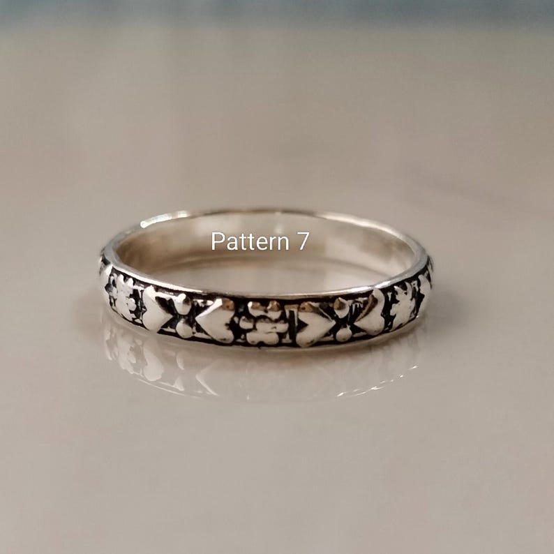 May include: A silver-toned metal ring featuring a repeating embossed pattern of hearts and small floral shapes around the band. The text Pattern 7 is printed in white in the centre of the ring. The metal has a polished finish with dark accents.