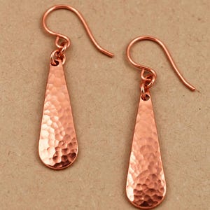 May include: A pair of copper drop earrings featuring a teardrop shape with a hammered, textured finish. Each earring is suspended from a simple curved ear wire hook, showcasing the metallic reddish-brown color and reflective surface of the metal.