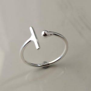 Peut inclure: Une bague ouverte de couleur argentée présentant une barre en forme de T à une extrémité et une petite perle sphérique à l'autre. L'anneau est fin et circulaire, conçu avec un espace réglable entre les deux extrémités au fini métallique poli.
