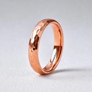 Op de afbeelding: Een roségouden ring met een gehamerd, getextureerd oppervlak. De ring heeft een afgerond profiel en een gepolijste afwerking die het licht reflecteert over de onregelmatige, gefacetteerde buitenkant. De binnenkant is glad en bevat de tekst TheHeavenOfCraft.
