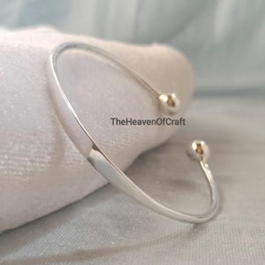 May include: A polished silver-tone open cuff bracelet featuring a smooth, curved band that terminates in two small, rounded ball ends. The text TheHeavenOfCraft is printed in black lettering across the centre of the metallic surface.