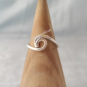 Puede incluir: Un anillo de plata con un diseño de onda continua que se curva en una espiral central. El metal tiene un acabado pulido y reflectante. El texto TheHeavenOfCraft es visible en letras blancas sobre el centro de la banda del anillo.