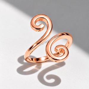 May include: A rose gold-toned ring featuring an open, bypass-style design with two prominent, symmetrical spiral ends. The metal surface is smooth and polished, reflecting light. The text TheHeavenOfCraft is visible on the ring band.