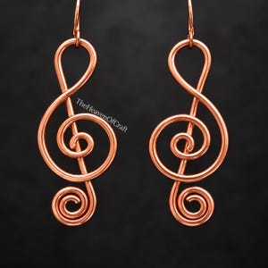 May include: A pair of copper-colored wire earrings shaped like treble clef musical symbols. Each earring features a continuous looped wire design with a hook at the top. The text TheHeavenOfCraft is visible in white lettering across the left earring.