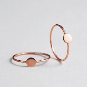 May include: Two thin rose gold rings, each featuring a small, flat, circular disc element. The metal bands have a subtle hammered texture, and the text TheHeavenOfCraft is visible in small black lettering near the centre of the horizontal ring.