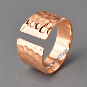 Könnte beinhalten: Ein breiter, offener Ring aus poliertem roségoldfarbenem Metall. Die Außenfläche weist eine ausgeprägte gehämmerte Textur mit zahlreichen kleinen, abgerundeten Vertiefungen auf. Der Text TheHeavenOfCraft ist an der Seite des Rings aufgedruckt.