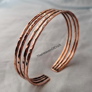 May include: A copper cuff bracelet featuring four parallel bands with a hammered, textured finish. The metal has a warm, reddish-brown metallic luster. The text TheHeavenOfCraft is visible in black lettering on the surface of the bracelet.