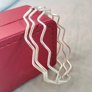 May include: Two silver-toned bangles featuring a continuous wavy, zigzag pattern. The bracelets are constructed from thin, polished metal wire formed into a repeating undulating shape, creating a stackable set of circular wrist jewellery.