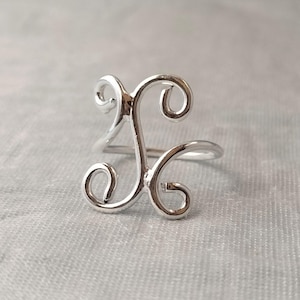 Könnte beinhalten: Ein silberfarbener Ring mit einem dekorativen, stilisierten Buchstaben K als Herzstück. Der Buchstabe besteht aus glattem, poliertem Metalldraht, der sich an den oberen und unteren Ecken zu vier ausgeprägten Schlaufen biegt und ein offenes Design bildet.