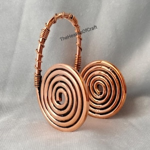 Op de afbeelding: Een koperdraad accessoire met twee platte cirkelvormige spiralen verbonden door een gebogen, met draad omwikkelde boog. Het metaal heeft een gepolijste, warme roodbruine afwerking. De tekst TheHeavenOfCraft is in zwarte letters op de boog zichtbaar.
