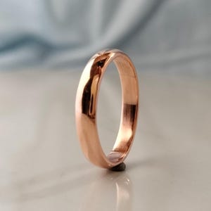 May include: A polished rose gold ring with a smooth, rounded band. The metal surface features a high-shine finish that reflects light, creating bright highlights along the curved edges of the circular band, which stands upright on its edge.
