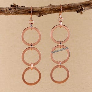 May include: A pair of rose gold-toned drop earrings, each featuring three vertically linked circular rings suspended from a hook. The text TheHeavenOfCraft is printed in small black letters on the right earring, hanging from a brown twig.