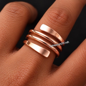 Puede incluir: Un anillo de metal color oro rosa con un diseño envolvente de varias capas con varias bandas paralelas que se enroscan alrededor del dedo. La superficie del metal es lisa y reflectante. El texto TheHeavenOfCraft es visible en el anillo.