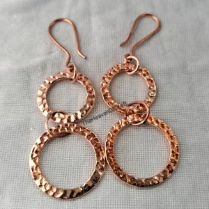 May include: A pair of copper dangle earrings featuring two interconnected hammered circles of varying sizes. Each earring is suspended from a curved ear wire. The metal surface has a distinct textured, dimpled finish that reflects light.