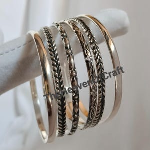 Puede incluir: Un conjunto de cinco brazaletes de metal plateado con texturas variadas. La colección incluye una banda lisa pulida, dos bandas de estilo trenzado, una banda con un patrón floral repetitivo y una banda con un diseño de cuentas y hojas.