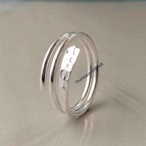 May include: A silver-coloured bypass ring featuring a double-band design. One band has a smooth, polished finish, while the other band displays a textured, hammered surface. The text TheHeavenOfCraft is printed in black on the inner side of the ring.