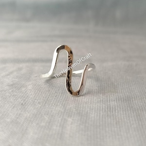 May include: A silver ring featuring a continuous, wavy, open-ended band with a hammered texture on the front surface. The text TheHeavenOfCraft is visible across the centre of the ring. The metal has a polished, reflective finish throughout.