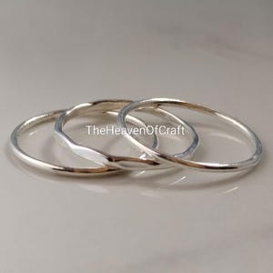 Può includere: Tre anelli sottili in metallo color argento disposti in una pila sovrapposta. Gli anelli hanno una finitura liscia e lucida con un profilo arrotondato. Il testo TheHeavenOfCraft è stampato al centro degli anelli in un carattere scuro.