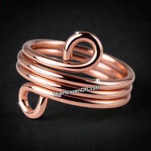 Puede incluir: Un anillo de metal color oro rosa con un diseño de alambre continuo que se envuelve sobre sí mismo en múltiples espirales, terminando en dos pequeños bucles. El texto TheHeavenOfCraft está impreso en negro sobre la superficie metálica.