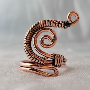May include: A copper wire ring featuring a coiled design with a smooth base band and an upper section composed of tightly wrapped wire spirals that curve upwards into a circular terminal point, showcasing a metallic reddish-brown finish throughout.
