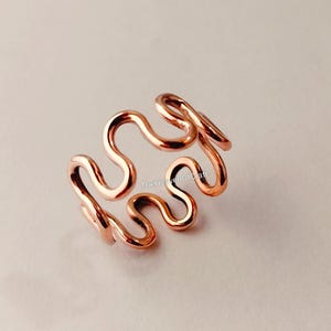 Puede incluir: Un anillo en tono oro rosa que presenta un diseño de alambre ondulado o serpentino continuo y repetitivo. El metal tiene un acabado pulido y reflectante. El texto TheHeavenOfCraft es visible en letras pequeñas y oscuras sobre el anillo.
