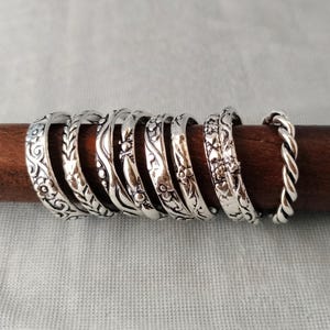 May include: A collection of eight silver-toned rings arranged in a row on a dark wooden rod. The rings feature various embossed patterns including floral motifs, leaf designs, wavy lines, small dots, heart shapes, and a twisted rope texture.
