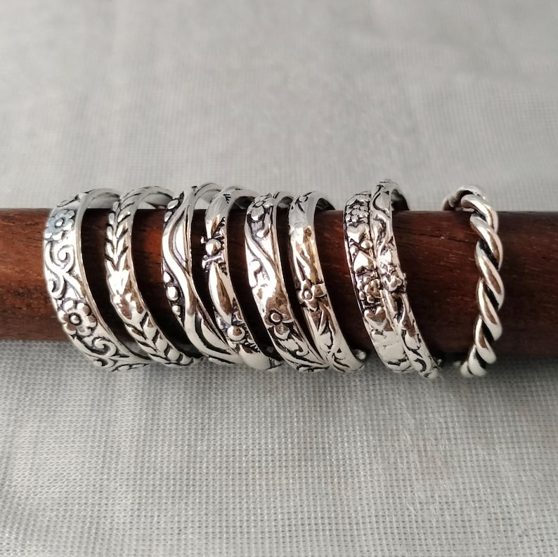 May include: A collection of eight silver-toned rings arranged in a row on a dark wooden rod. The rings feature various embossed patterns including floral motifs, leaf designs, wavy lines, small dots, heart shapes, and a twisted rope texture.