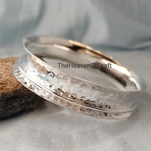 Puede incluir: Un brazalete de color plateado que presenta una banda ancha, martillada y texturizada con un borde interior liso y pulido. La superficie metálica refleja la luz, creando brillos intensos en la forma curva. El texto TheHeavenOfCraft es visible.