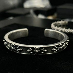 Könnte beinhalten: Silberfarbenes Armreif-Armband mit floralem Design. Der offene Armreif hat eine Reihe stilisierter Blumenmotive um das Band. Das Armband ist vor einem dunklen Hintergrund platziert, was seinen metallischen Glanz hervorhebt.
