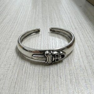 May include: A silver cuff bracelet featuring a decorative dagger and floral design. The open-ended bracelet is made of shiny metal. The dagger has a detailed handle and is centered on the bracelet.