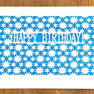 May include: A white card with a blue patterned background. The pattern is made up of stars and diamonds. The words "HAPPY BIRTHDAY" are cut out of the card.