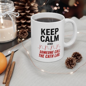May include: A white ceramic mug featuring the black text KEEP CALM AND, a red EKG heart rhythm line, and the red text SOMEONE CALL THE CATH LAB. The mug has a standard handle and a smooth, cylindrical body with a glossy finish.