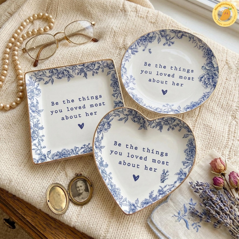 May include: Three decorative white ceramic dishes with blue floral patterns and gold trim. The dishes are square, round, and heart-shaped, each bearing the inscription "Be the things you loved most about her." A vintage locket and eyeglasses are also present.