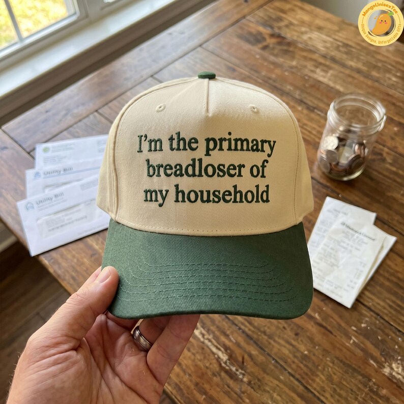 K&ouml;nnte beinhalten: Beige und gr&uuml;ne Baseballkappe mit dem Text "I'm the primary breadloser of my household" auf der Vorderseite gestickt. Die Kappe hat einen gr&uuml;nen Schirm und einen kleinen gr&uuml;nen Knopf oben. Die Kappe wird in einer Hand gehalten.
