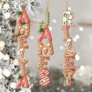 May include: Gingerbread-style Christmas ornaments spelling out the names AMELIA, JAMES, and RILEY. Each letter is decorated with icing, sprinkles, and holly. The ornaments hang from gold strings against a blurred, snowy background.