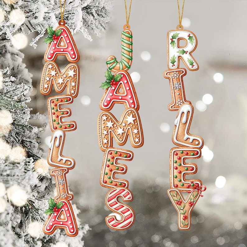 May include: Gingerbread-style Christmas ornaments spelling out the names AMELIA, JAMES, and RILEY. Each letter is decorated with icing, sprinkles, and holly. The ornaments hang from gold strings against a blurred, snowy background.