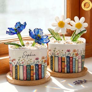 May include: Two white ceramic flower pots with colorful floral and book designs. One pot has blue glass flowers, the other has white glass daisies. The name "Sophia" is written on one pot. Both pots sit on wooden coasters.