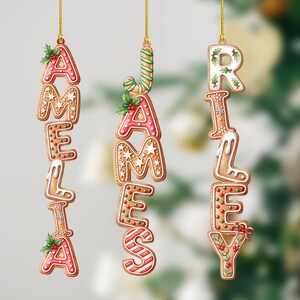 May include: Gingerbread-style Christmas ornaments spelling out the names "Amelia", "James", and "Riley". Each letter is decorated with icing, holly, and other festive details, suspended by gold strings.