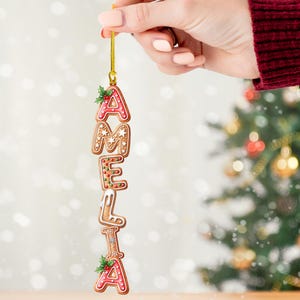 May include: A Christmas ornament spelling "AMELIA" in gingerbread-style letters. Each letter is decorated with red, white, and green icing, and holly accents. The ornament hangs from a gold string, perfect for holiday decor.