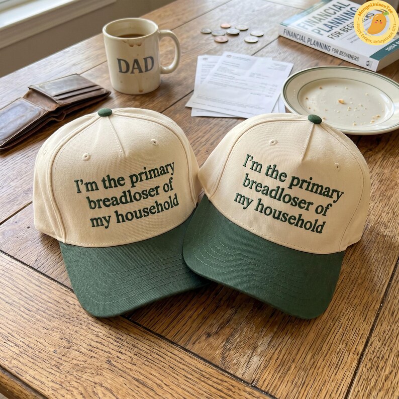 K&ouml;nnte beinhalten: Zwei beige Baseballkappen mit gr&uuml;nen Schirmen liegen auf einem Holztisch. Auf jeder Kappe steht in schwarzer Schrift "I'm the primary breadloser of my household". Eine Tasse mit der Aufschrift "DAD" ist ebenfalls zu sehen.