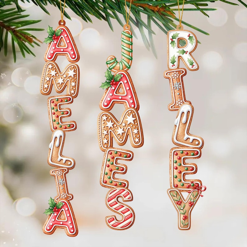 May include: Christmas ornaments spelling out the names AMELIA, JAMES, and RILEY. The ornaments are designed to look like gingerbread cookies, with red, white, and green icing and decorative details. They hang from a pine branch.